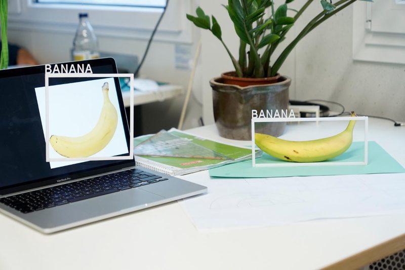 A banana on a table and an image of a banana on a laptop on the same table. Each of the two bananas has a white frame around it with the word ‚Banana‘ sticked on top of it.