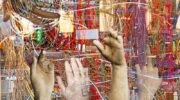 The image depicts a series of stone-like hands, fractured and cracked, reaching out in various directions from a chaotic mass of tangled wires. Some hands display broken fingers, while archival tags on others suggest anonymity and erasure. The juxtaposition of human forms and technological wires evokes themes of labor, exploitation, and the often unseen physical work behind the tech industry.