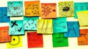 Seventeen multicoloured post-it notes are roughly positioned in a strip shape on a white board. Each one of them has a hand drawn sketch in pen on them, answering the prompt on one of the post-it notes "AI is...." The sketches are all very different, some are patterns representing data, some are cartoons, some show drawings of things like data centres, or stick figure drawings of the people involved.
